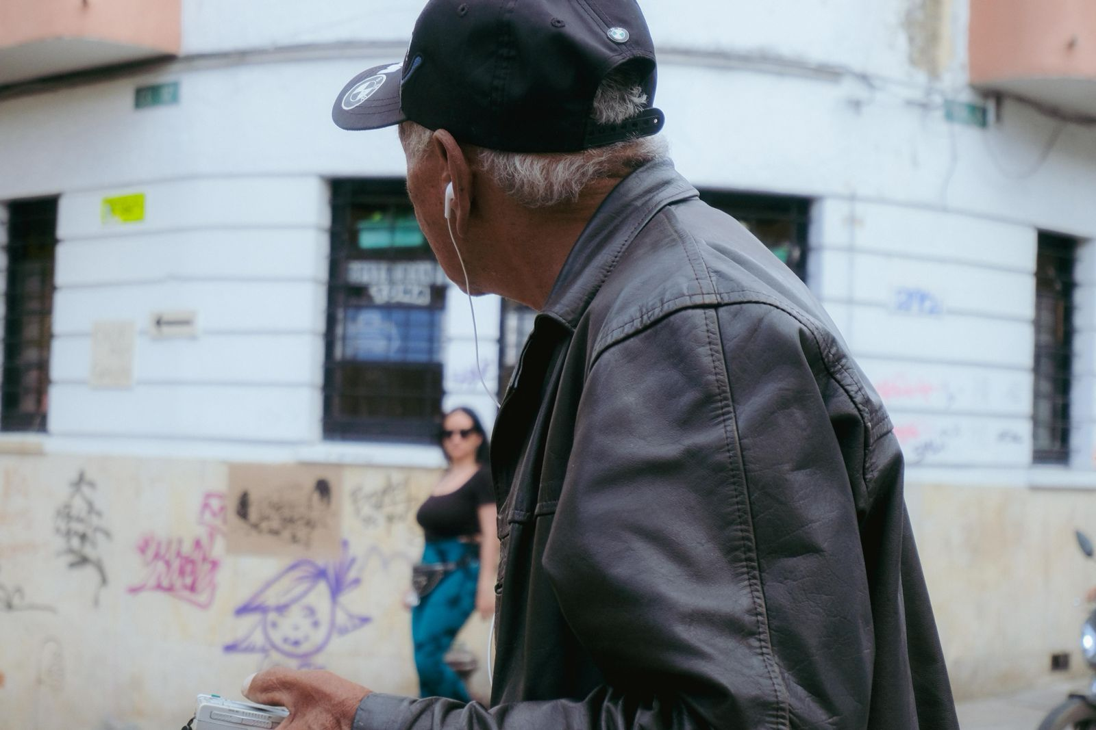 Homem idoso de boné preto e jaqueta cinza, de costas, em uma rua urbana com grafites ao fundo