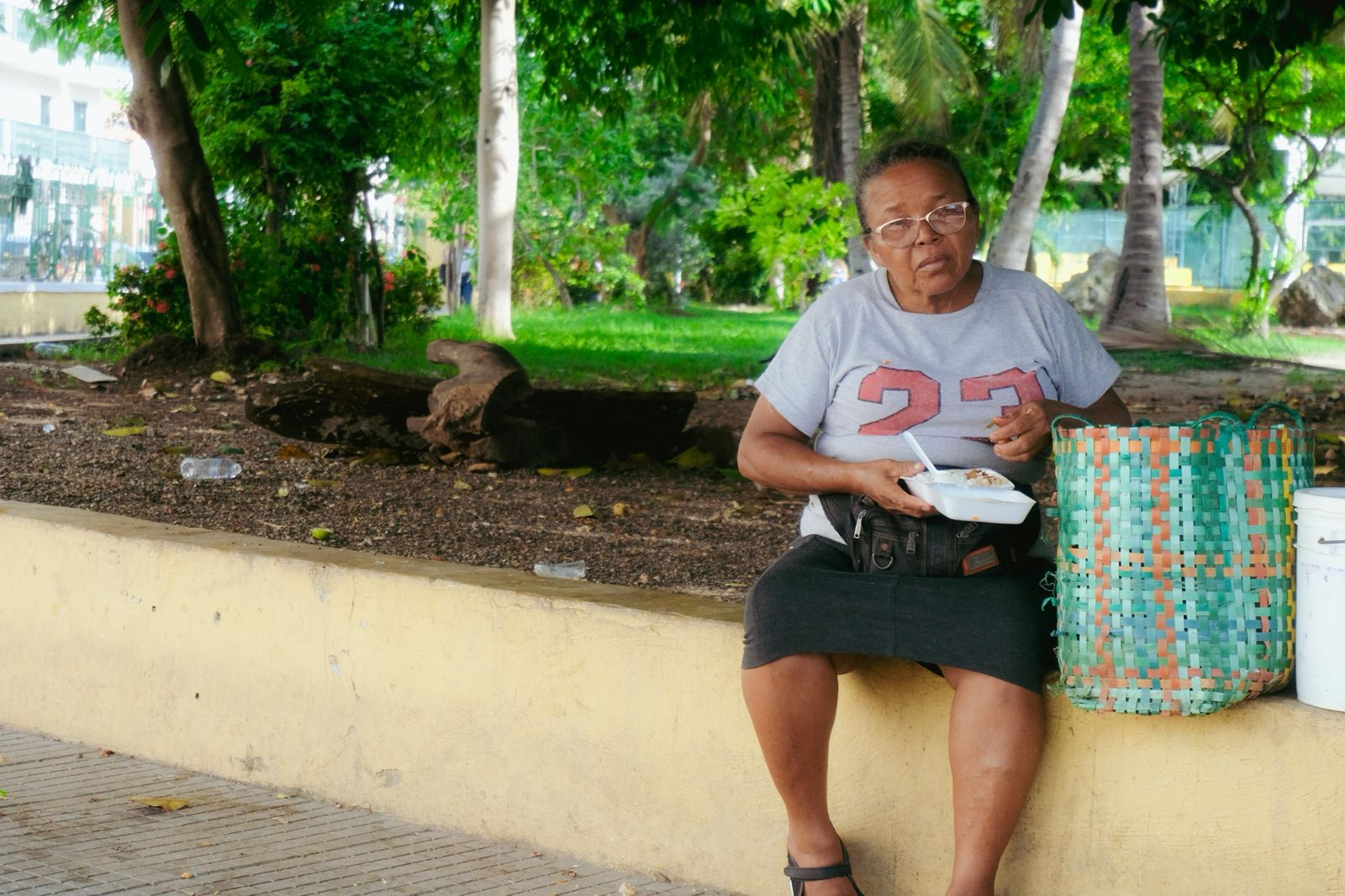 Mulher sentada em um muro ao ar livre, comendo de uma marmita, com uma cesta trançada ao lado e árvores ao fundo